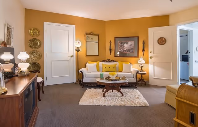 A cozy living room with mustard yellow walls, a white sofa with yellow and white cushions, a round wooden coffee table on a white shag rug, two white paneled doors, vintage-style lamps, framed artwork, and decorative plates on the wall.