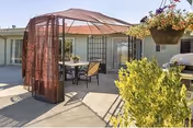 Sunlit outdoor courtyard with a fabric gazebo covering a table and chairs in front of a single-story building and hanging plants.