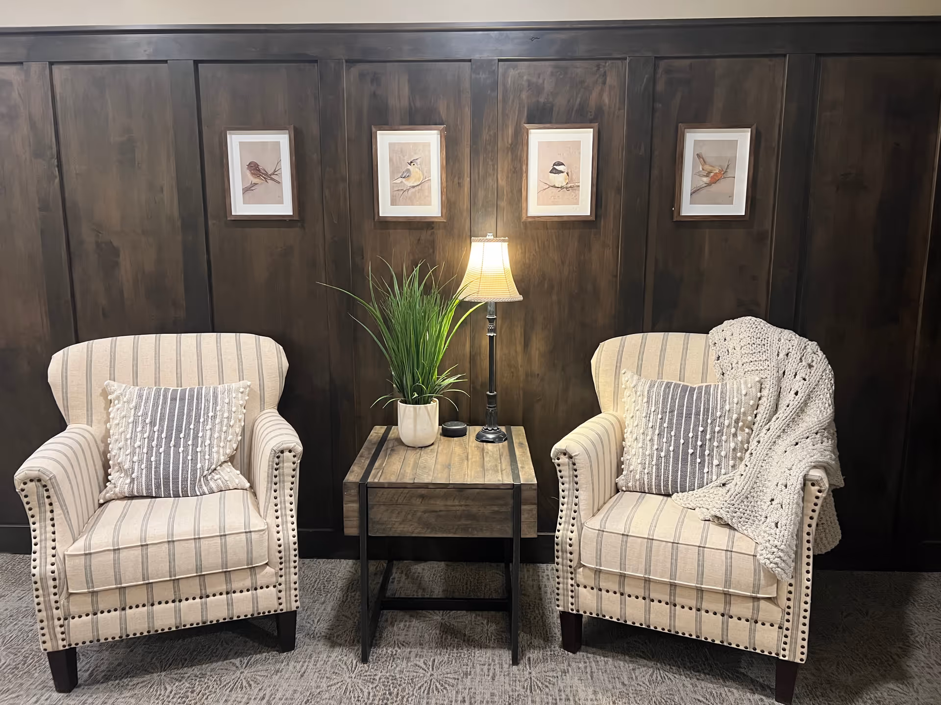 Two beige striped armchairs with decorative pillows and a knitted throw blanket on the right chair, positioned on either side of a wooden side table with a potted plant, a small lamp, and a smart speaker. The background features dark wood paneling with four framed bird illustrations hanging above.