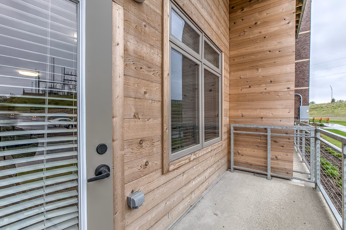 A small outdoor balcony area with wooden panel walls, a metal railing, and a glass door with blinds. The balcony overlooks a grassy area and a sidewalk, with a brick building visible on the right side.