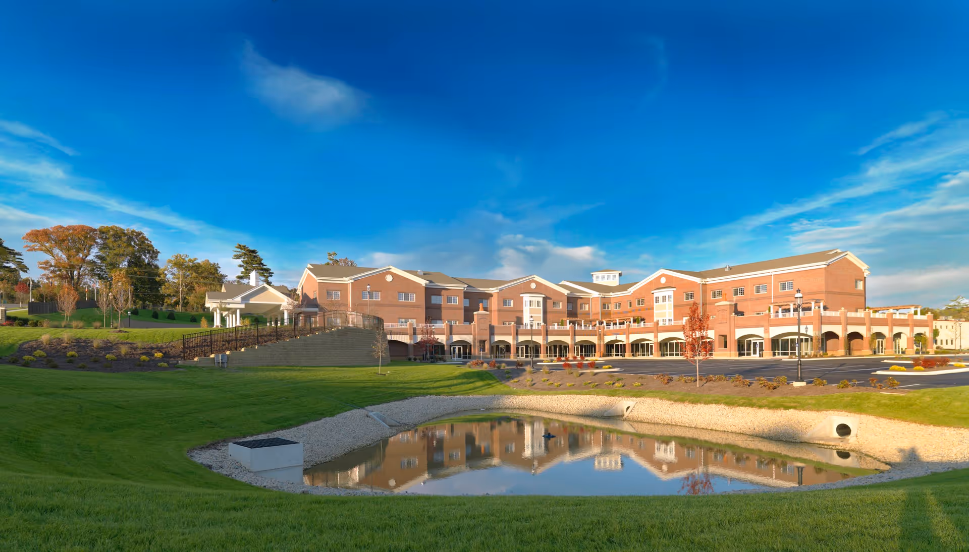 Large brick senior living building with manicured lawns and a small reflective pond under a blue sky.