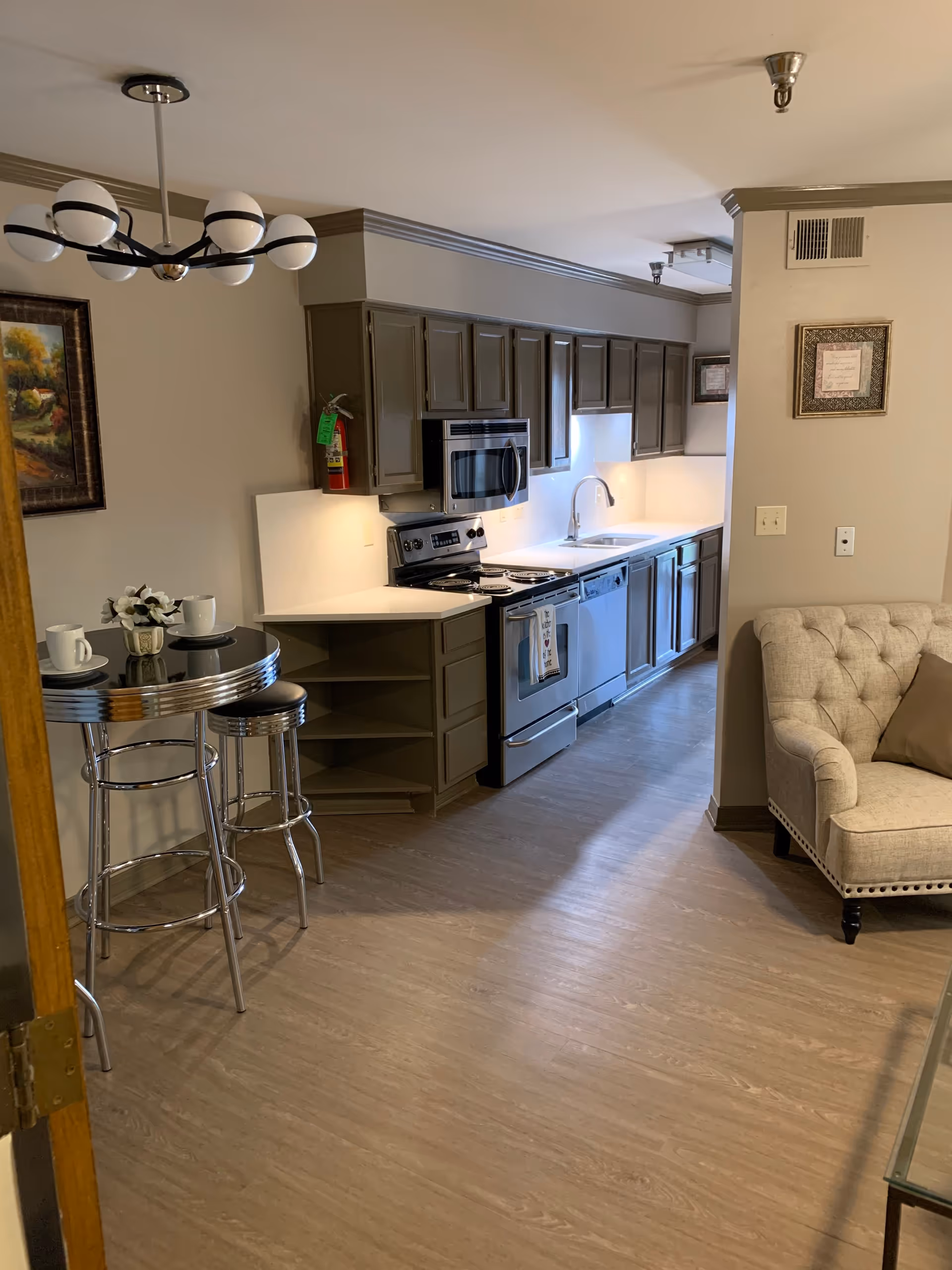 A modern kitchen area with gray cabinets, stainless steel appliances including a stove, microwave, and dishwasher. There is a small round table with two metal stools and a vase with white flowers. To the right, part of a beige tufted armchair is visible. The floor is light wood, and the walls are painted in neutral tones with a framed picture hanging on the wall.