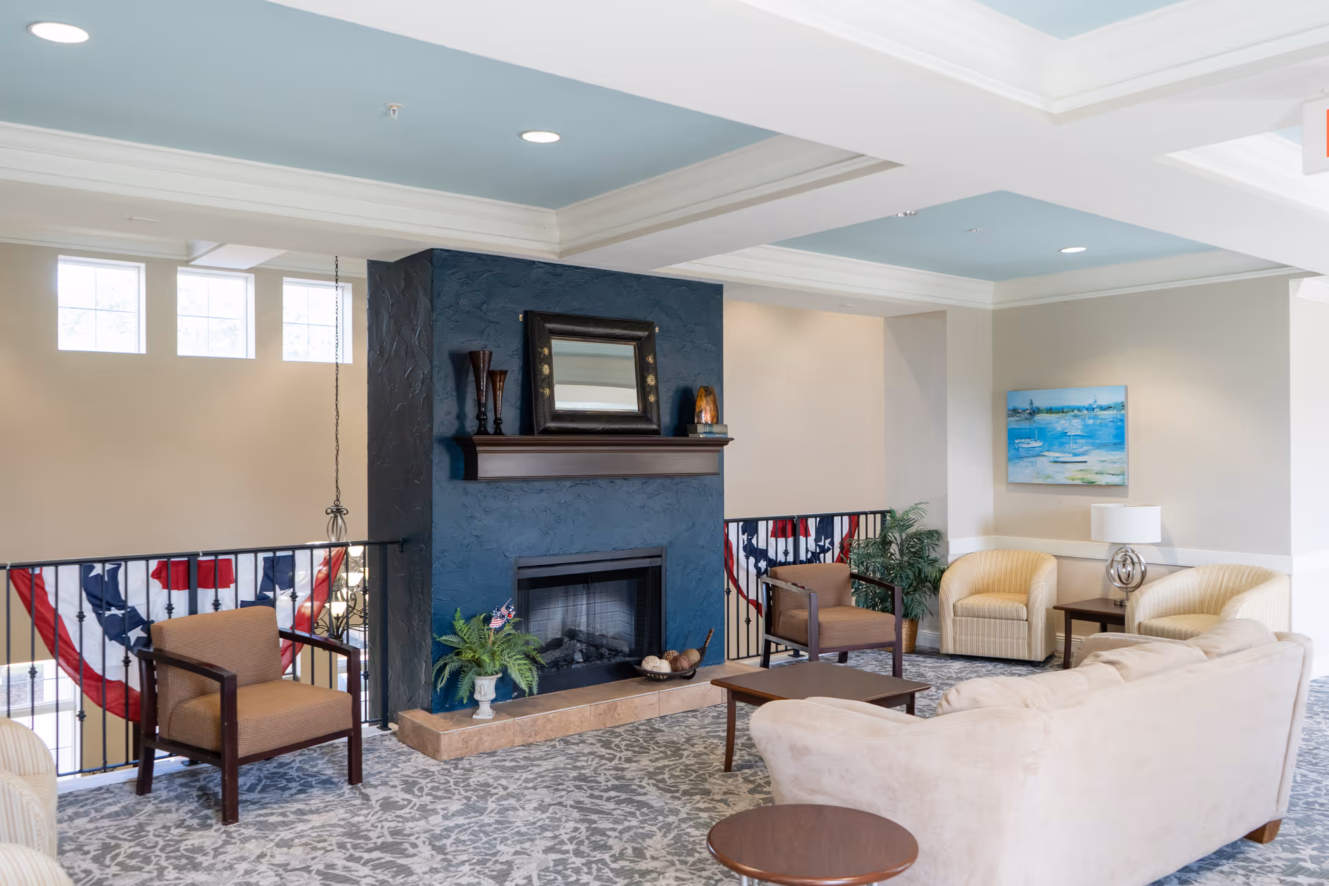 A cozy living room area with a beige sofa, two beige armchairs, and two brown wooden chairs arranged around a dark blue fireplace with a wooden mantel. The mantel holds decorative items including a mirror and vases. The room has light-colored walls, a patterned carpet, a painting on the wall, and a table lamp on a side table. There are windows near the ceiling letting in natural light and patriotic bunting hanging on the railing behind the chairs.