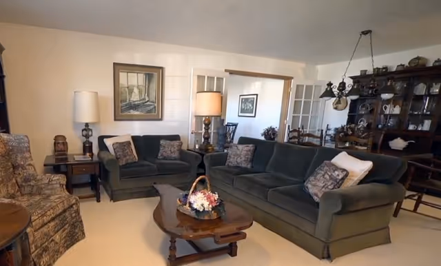 A cozy living room with two dark green sofas, a patterned armchair, a wooden coffee table with a flower basket centerpiece, two table lamps, and a large wooden cabinet with glass doors displaying decorative items. The room has light-colored walls and carpet, with framed artwork on the walls.