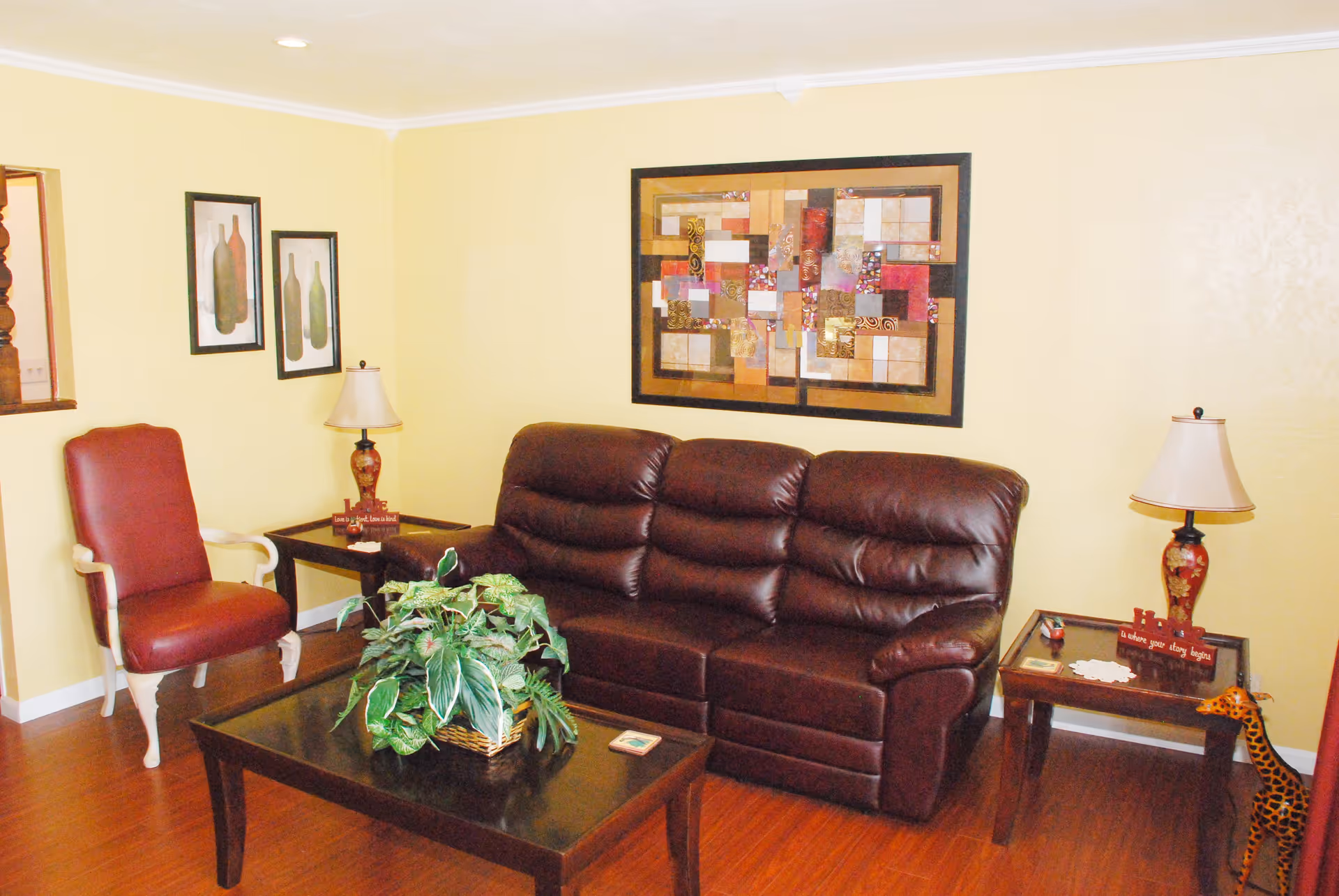 A cozy living room with a dark brown leather sofa, a red upholstered armchair, two side tables each with a decorative lamp, a coffee table with a green leafy plant in a basket, and framed artwork on yellow walls.