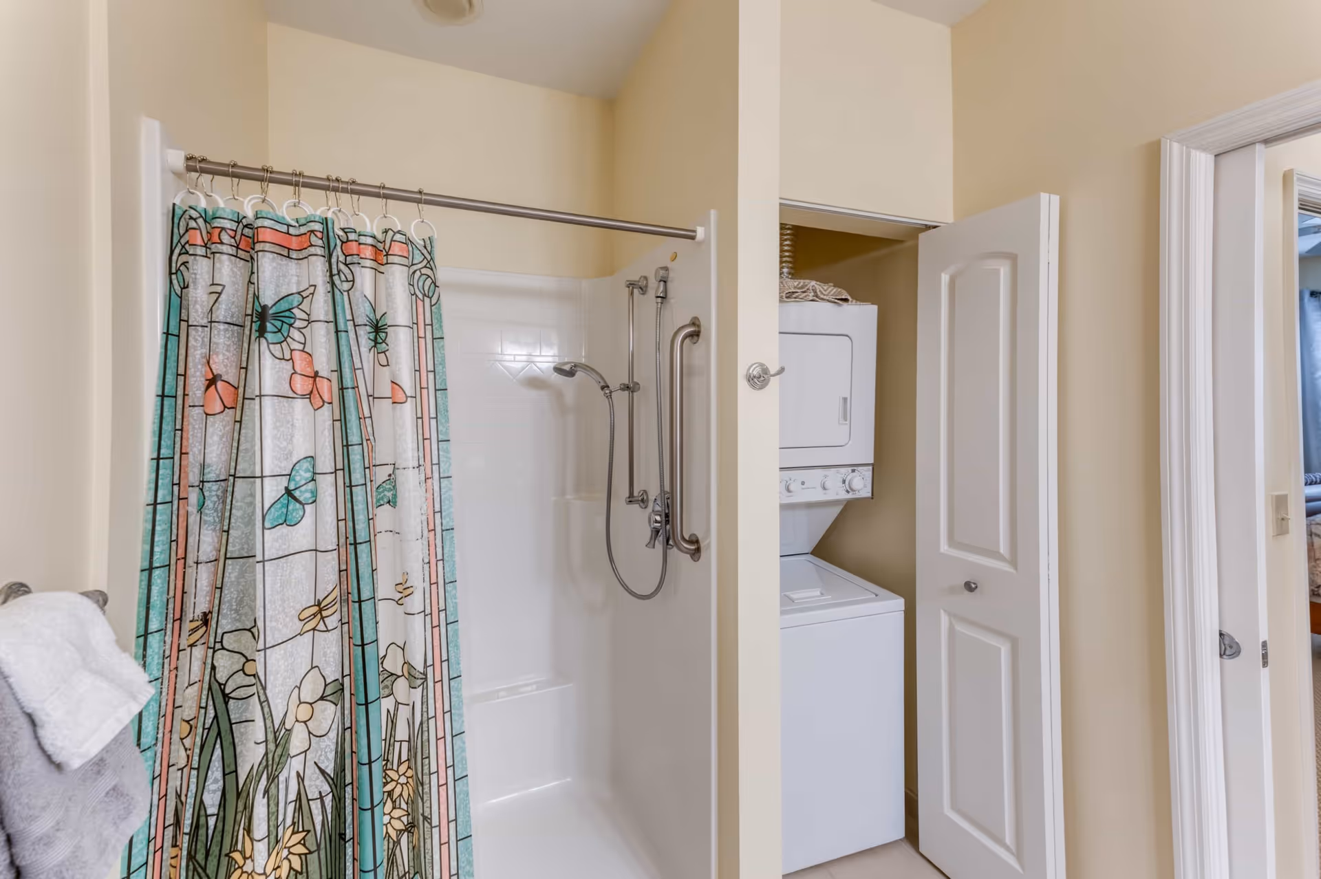 Bathroom with a shower area featuring a colorful shower curtain with butterfly and flower designs. Next to the shower is a small laundry closet with a stacked washer and dryer. The walls are painted beige and there is a towel hanging on a rack.
