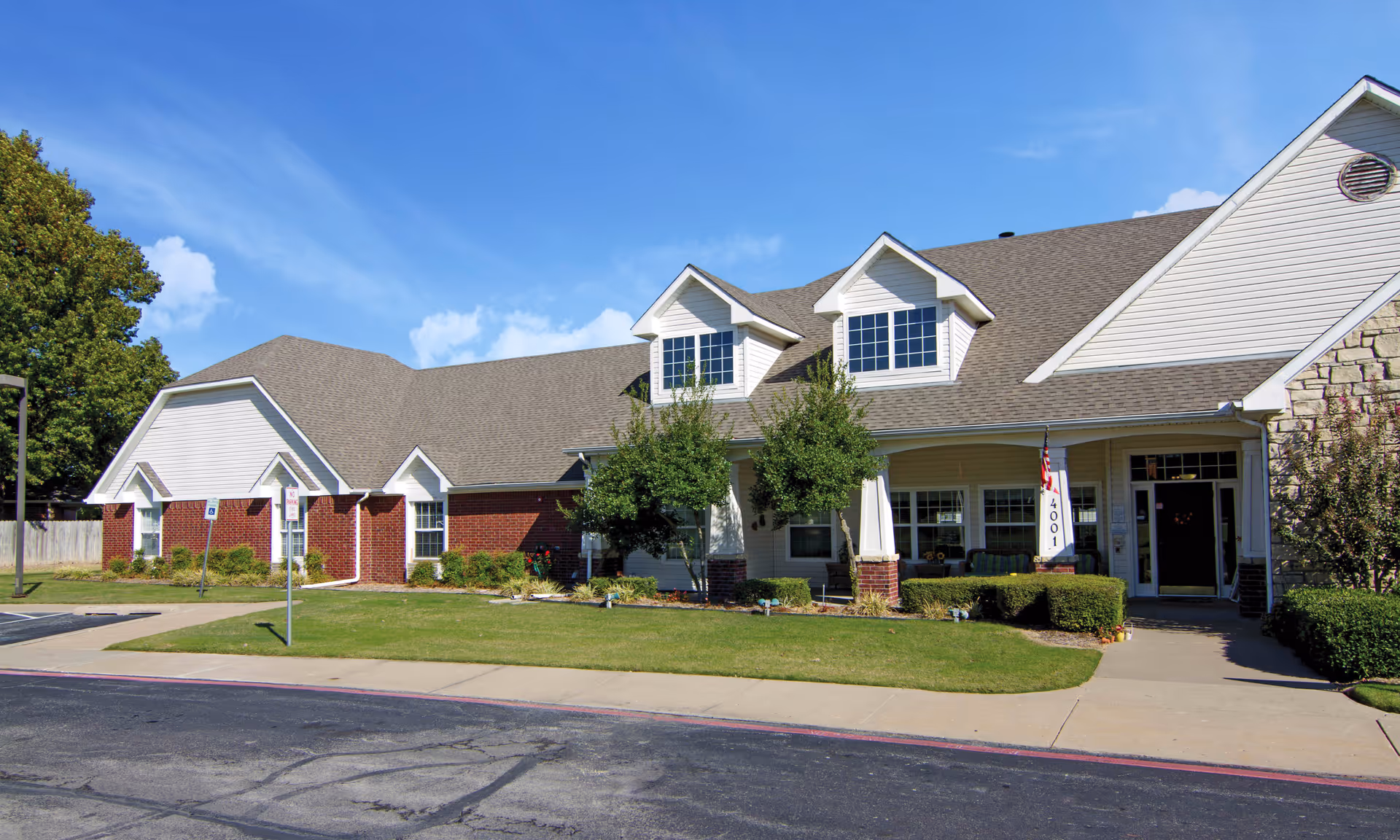 Front exterior of a single-story brick and siding senior living building with dormer windows, a covered porch, and a lawn under a blue sky.