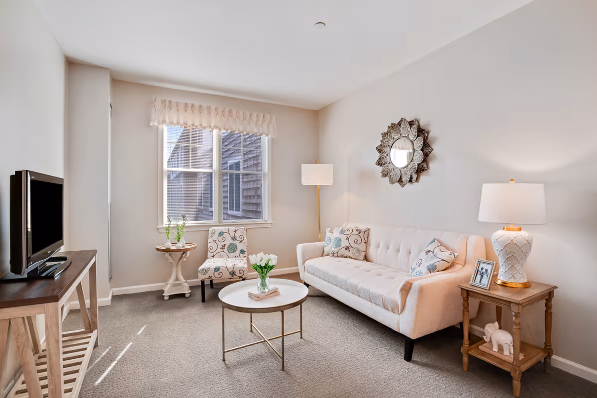 A cozy living room with a beige tufted sofa adorned with patterned pillows, a round white coffee table with a vase of white tulips, a floral upholstered chair, a wooden side table with a white lamp and a framed photo, and a flat-screen TV on a wooden stand. A decorative mirror hangs on the wall above the sofa, and a window with blinds and a valance lets in natural light.