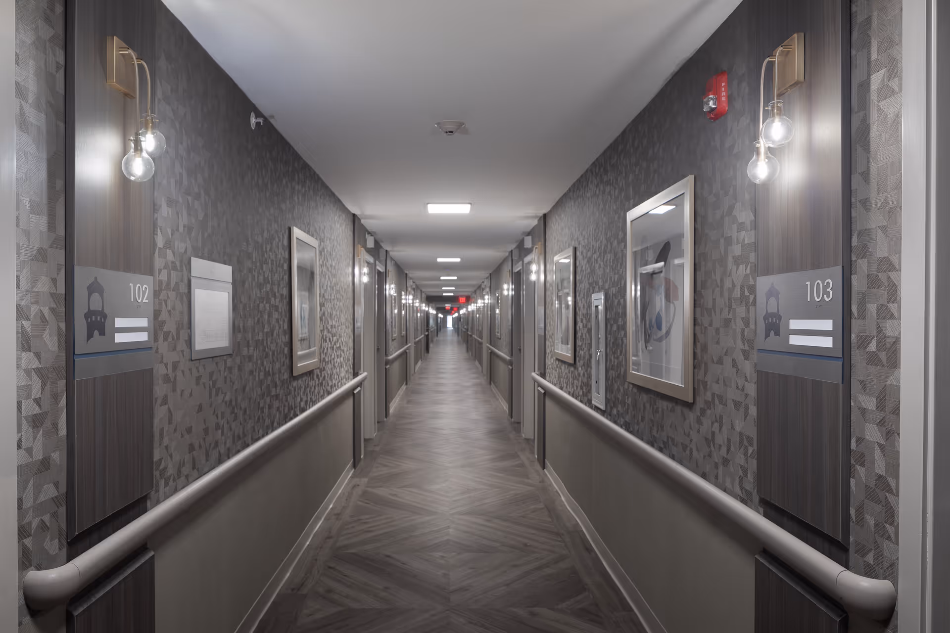 A long, well-lit hallway in a senior living facility with patterned wallpaper, handrails on both sides, framed artwork on the walls, and room number signs 102 and 103 visible near the foreground.