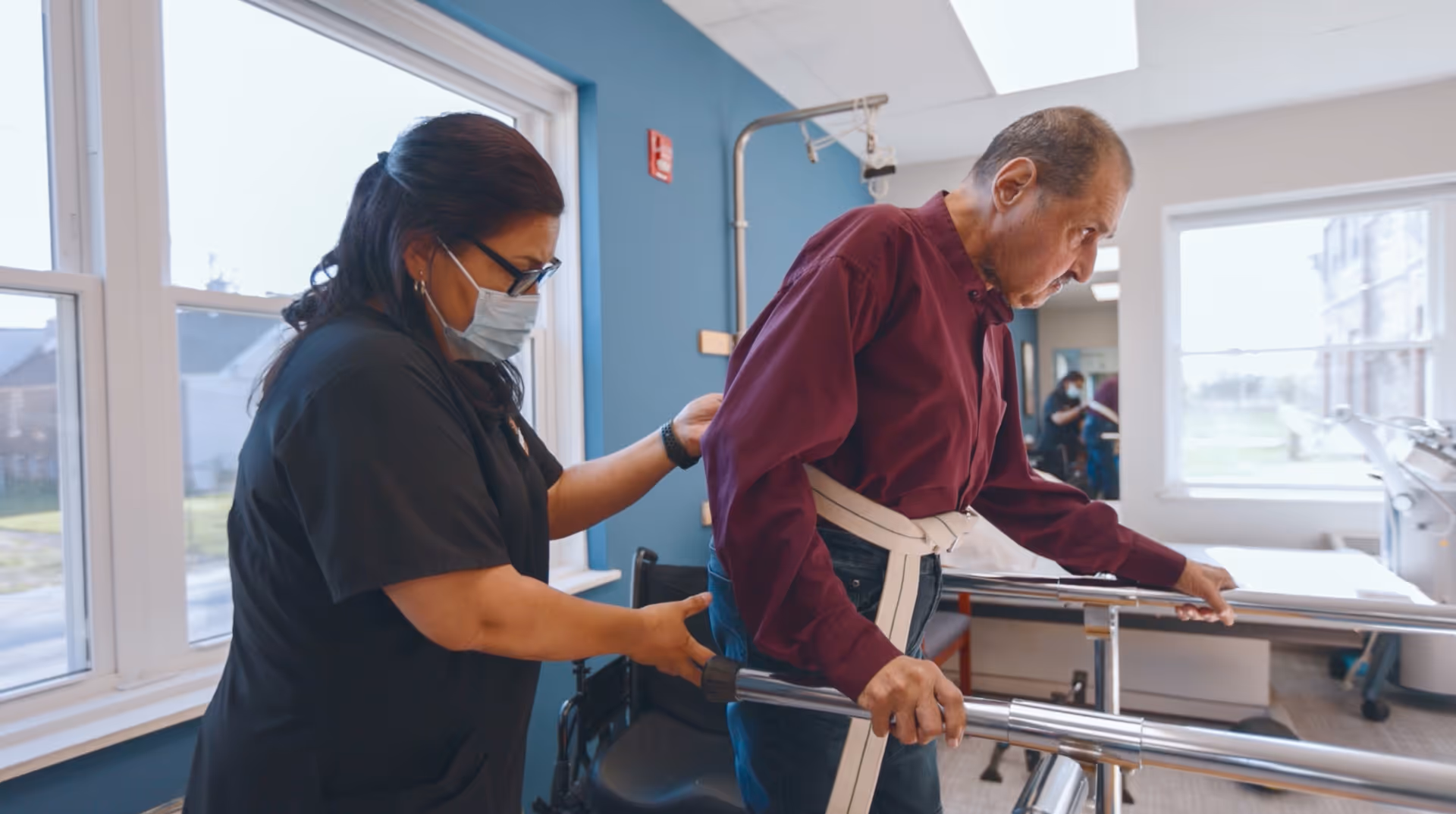 A healthcare worker wearing a mask assists an elderly man using a walker in a rehabilitation or therapy room with large windows and blue walls.