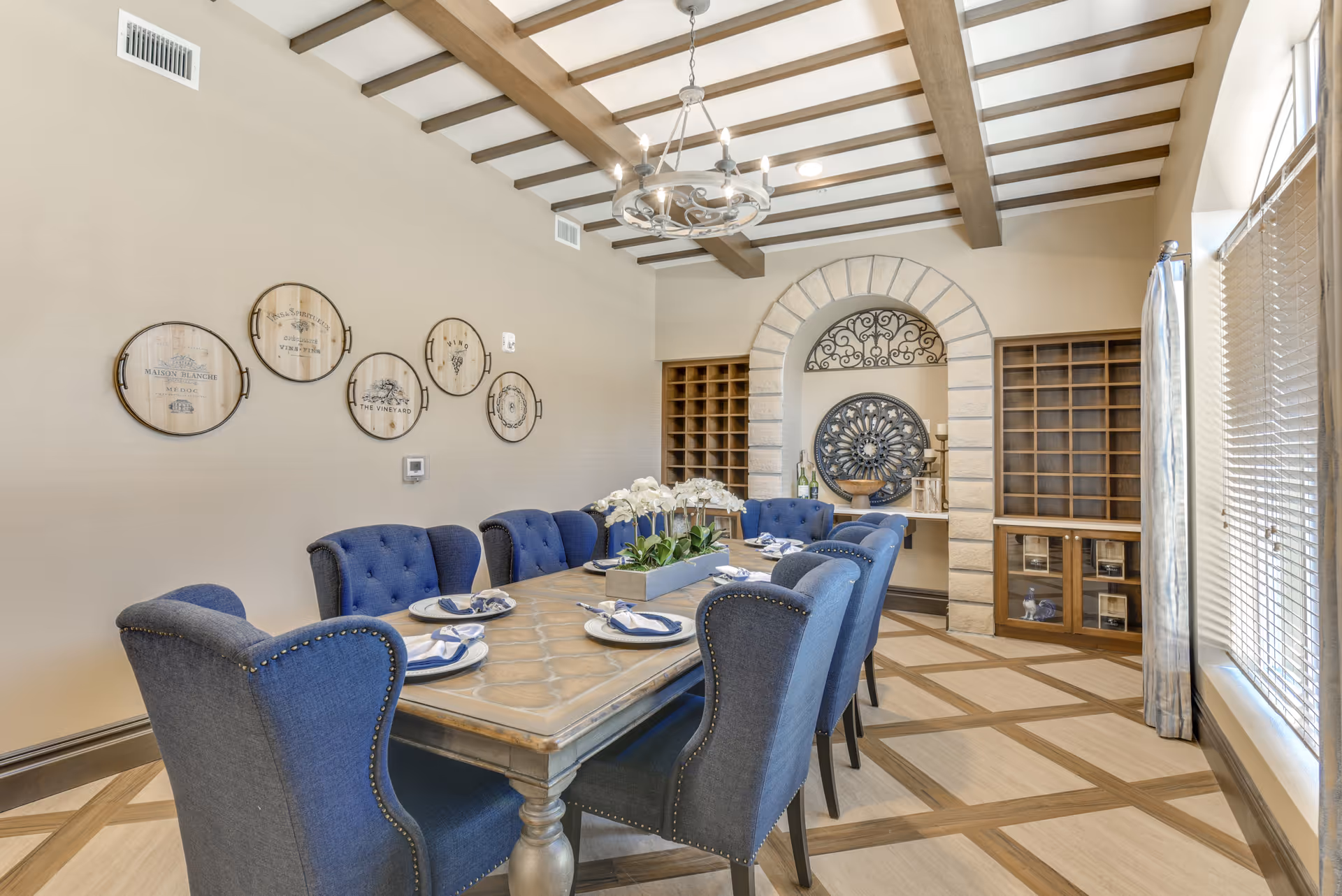 A dining room with a long wooden table set for six people with blue upholstered chairs. The room features a decorative chandelier, wooden ceiling beams, and wall art with circular designs. There are built-in wooden shelves and a stone archway with ornamental decor in the background. Large windows with blinds and curtains allow natural light into the room.