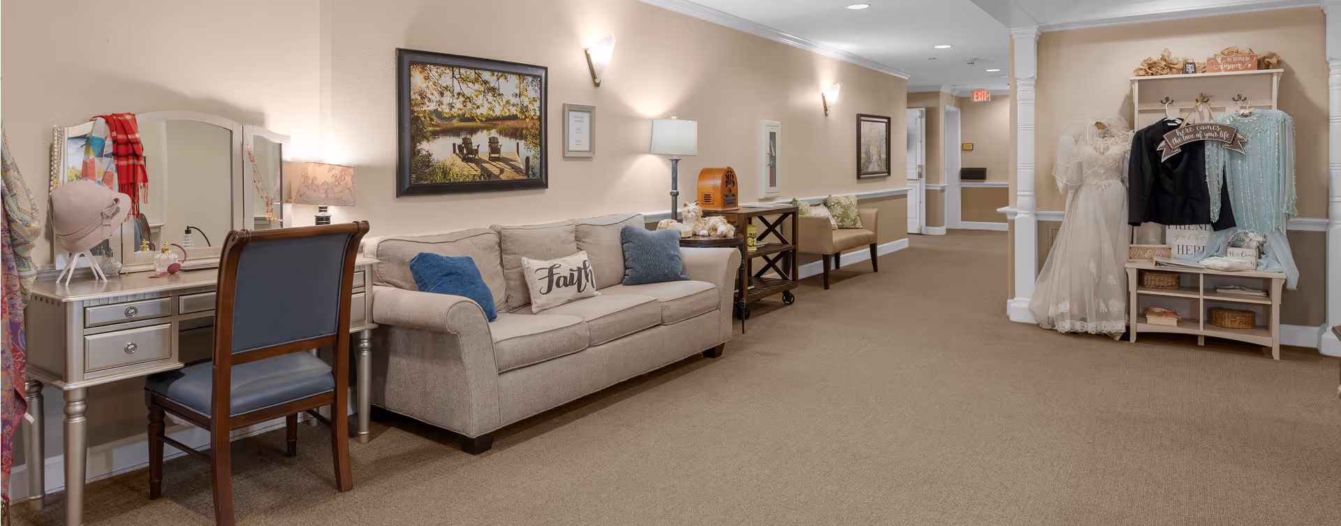 A cozy hallway area in a senior living facility featuring a beige sofa with decorative pillows, a silver vanity table with a chair and accessories, framed artwork on the walls, a small bench with cushions, and a display shelf holding wedding attire and decorative signs.