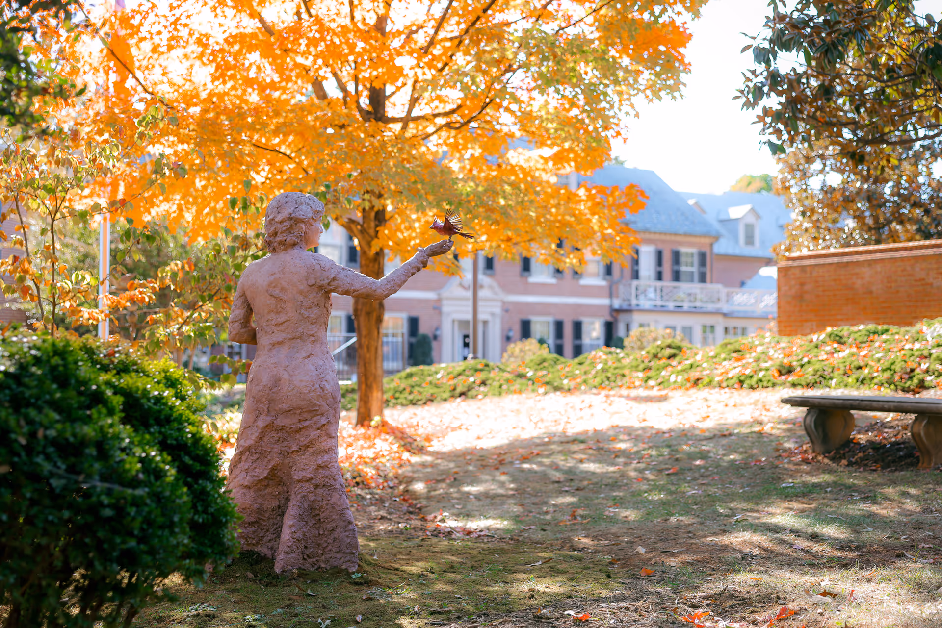 A garden courtyard showing a statue of a woman holding a bird amid autumn foliage with a brick building in the background.