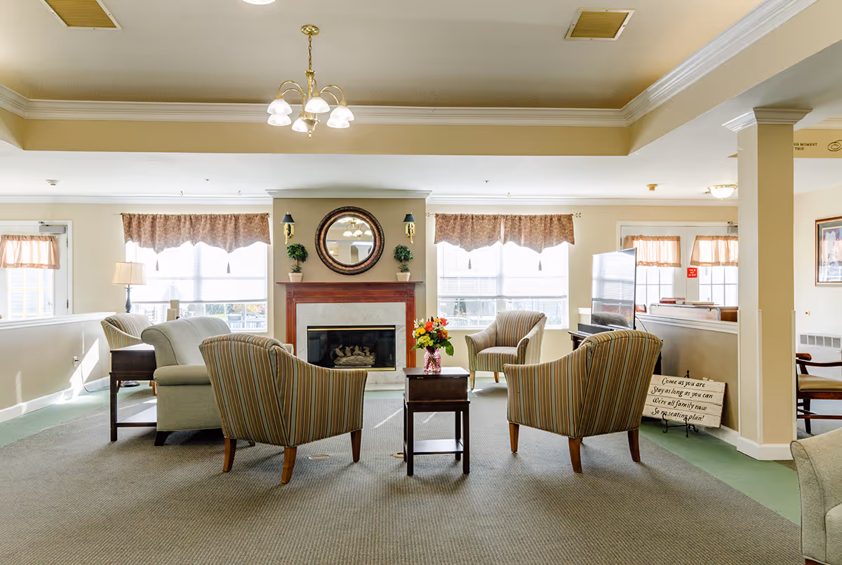 A bright and spacious living room area with a fireplace centered on the far wall, flanked by two windows with valance curtains. The room features several upholstered chairs and sofas arranged around a small wooden table with a vase of flowers. A round mirror and two small potted plants decorate the wall above the fireplace. The ceiling has a chandelier light fixture, and there is a television on the right side of the room. The overall decor is warm and inviting with beige walls and carpeted flooring.
