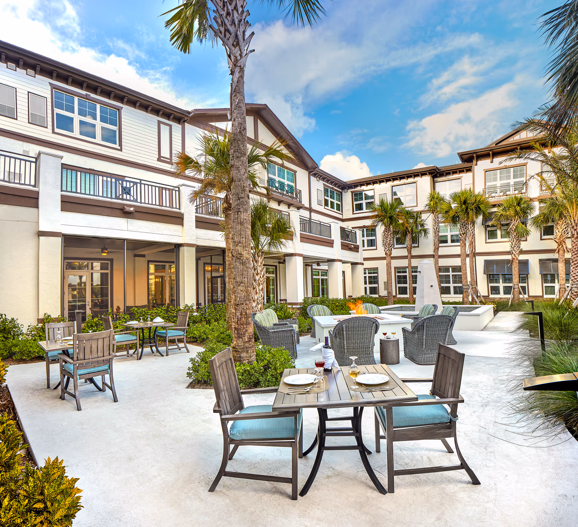 Outdoor patio area at Discovery Commons Hobe Sound featuring several tables with chairs, some set with plates and glasses. The patio is surrounded by palm trees and greenery, with a three-story building in the background under a partly cloudy blue sky.