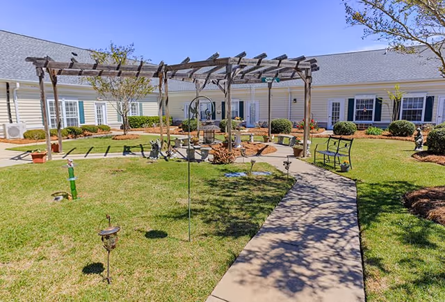 Outdoor courtyard area at Sumter Grove Senior Living featuring a wooden pergola, green lawn, paved walkway, benches, and landscaping with bushes and small trees in front of a single-story building with white siding and multiple windows.