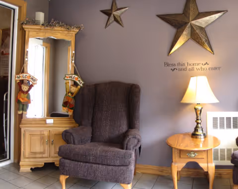 A cozy corner of a living room with a dark upholstered armchair, a wooden side table with a lit lamp, and a wooden cabinet with a mirror. The wall is decorated with two large metallic stars and a wall decal that reads 'Bless this home and all who enter'. Two small scarecrow decorations hang on the mirror.