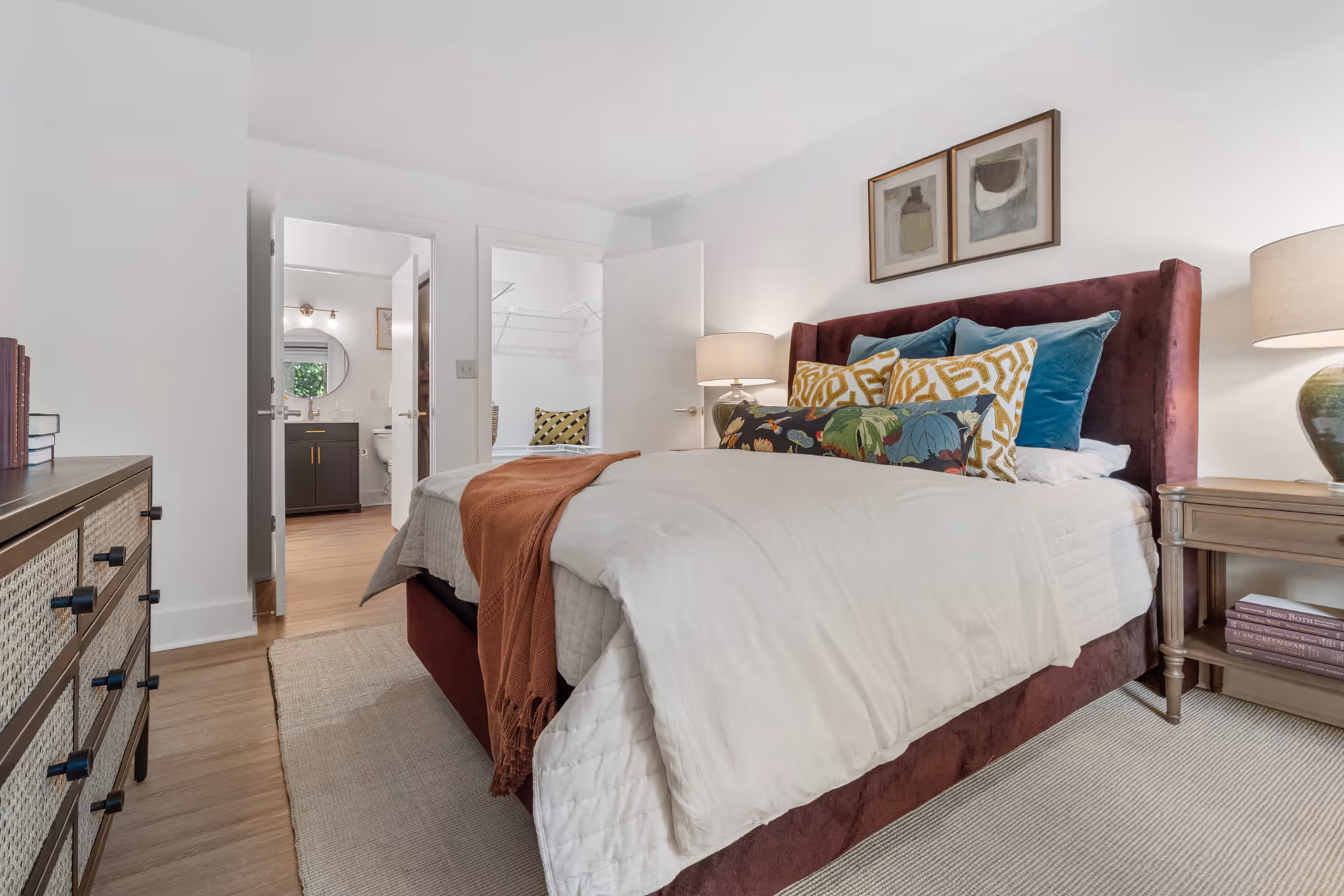 A cozy bedroom with a burgundy upholstered bed frame, white bedding, and multiple decorative pillows in blue, yellow, and floral patterns. There are two bedside tables with lamps on either side of the bed. On the left side of the image, there is a dresser with books on top. The room has light-colored walls and wooden flooring with a beige rug. Two open doors reveal a bathroom with a round mirror and a walk-in closet with a bench and pillows.