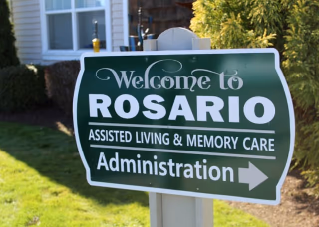 Green directional sign reading 'Welcome to ROSARIO Assisted Living & Memory Care Administration' posted on a lawn in front of a building.