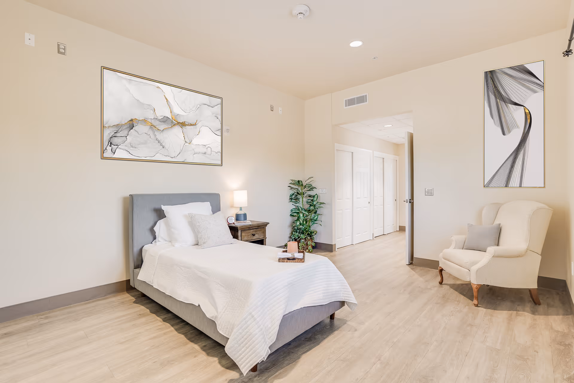 Bright, spacious single-occupancy bedroom with a bed, nightstand, armchair, and wall artwork.
