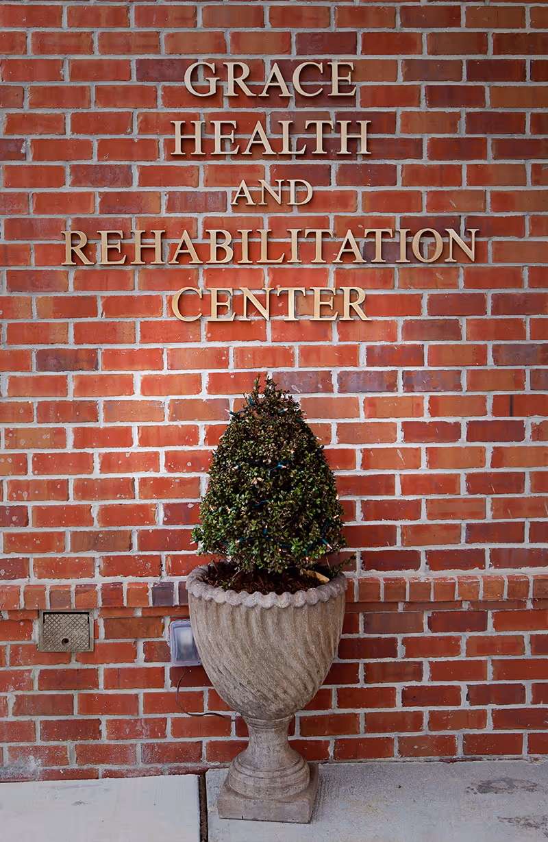 A brick wall with gold letters spelling out 'Grace Health and Rehabilitation Center' above a large decorative stone planter containing a small, trimmed shrub.