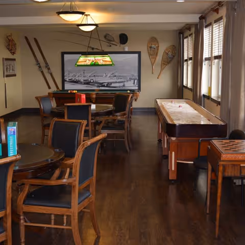 A recreational room with wooden floors and several tables and chairs. There is a pool table with a hanging light above it, a shuffleboard table, and a chess/checkers table. The walls are decorated with vintage skis, snowshoes, and a large black and white photo of airplanes. Windows with curtains line one side of the room.