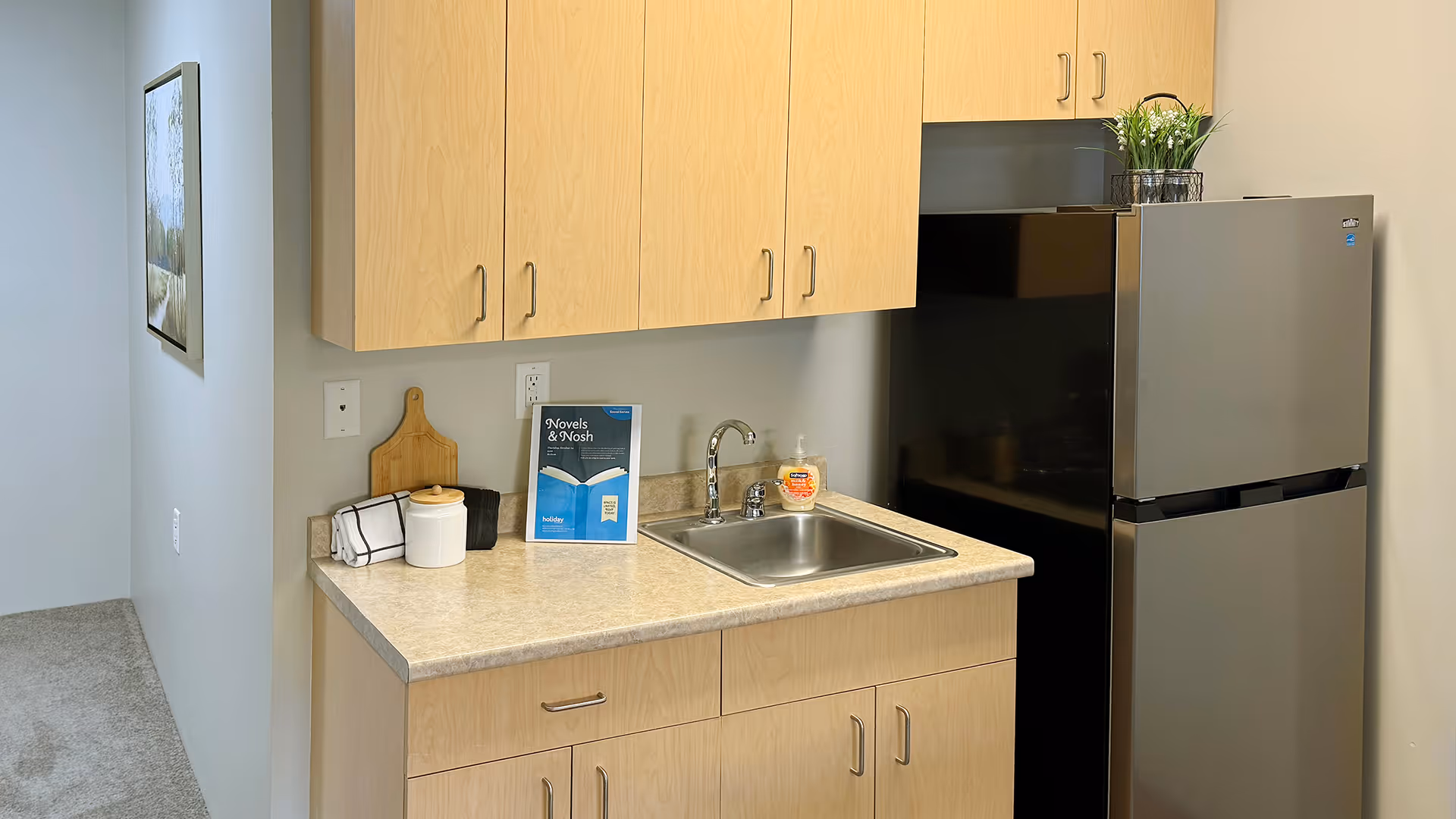 Small kitchenette with light wood cabinets, a countertop sink, and a stainless steel refrigerator.
