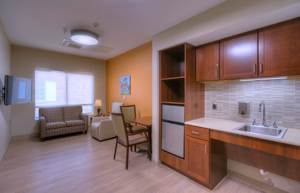 A cozy room in a retirement community featuring a small kitchenette with wooden cabinets, a sink, and a mini refrigerator. Adjacent to the kitchenette is a sitting area with a patterned loveseat, a beige armchair, a small table with two chairs, a lamp, and a wall-mounted flat-screen TV. The room has light wood flooring, a large window with blinds, and a warm orange accent wall with a framed picture.