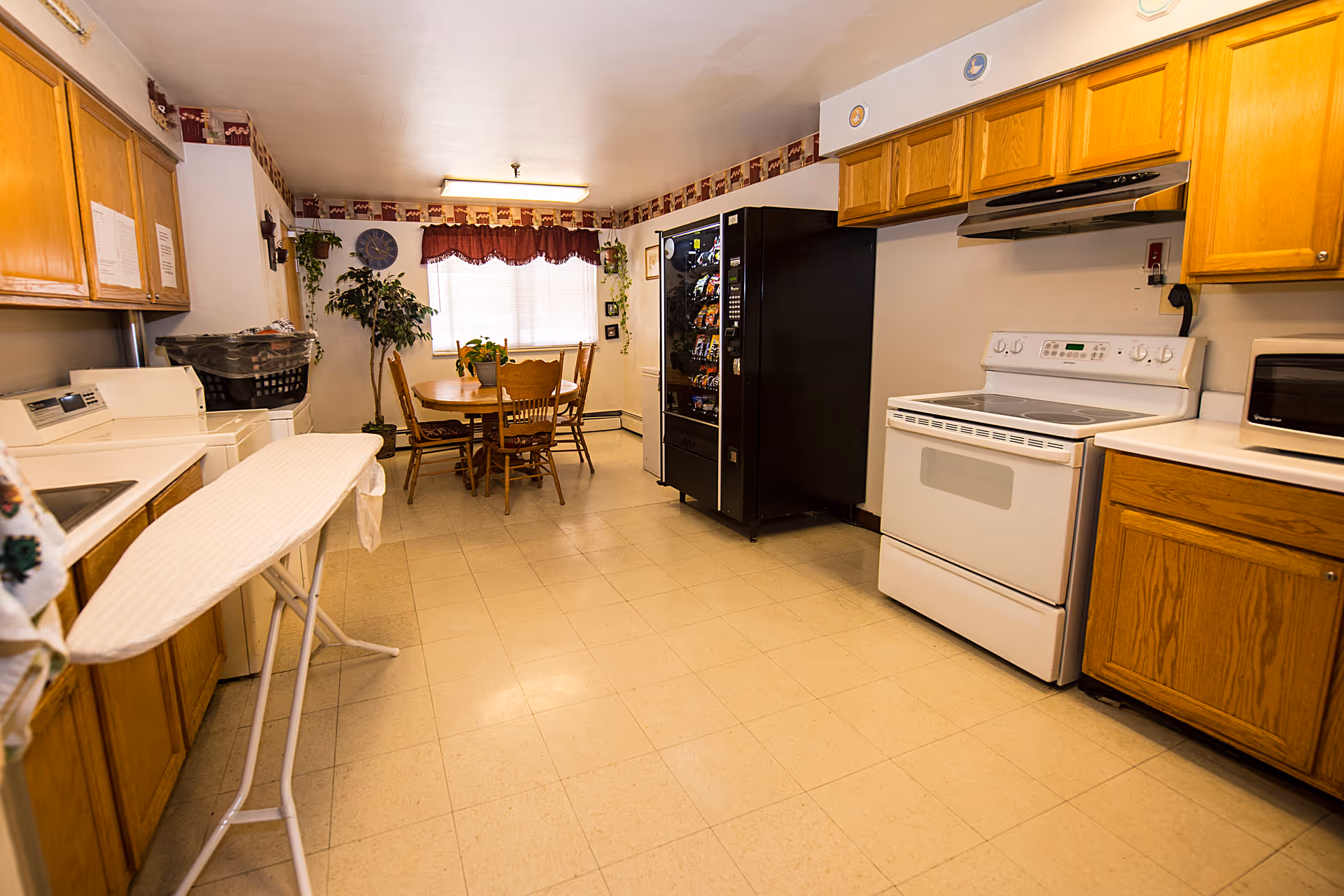A kitchen area with wooden cabinets, a white electric stove, microwave, and a black vending machine. There is an ironing board set up on the left side near a washing machine and dryer. At the far end, a round wooden dining table with four chairs is placed near a window with red curtains and plants around it.