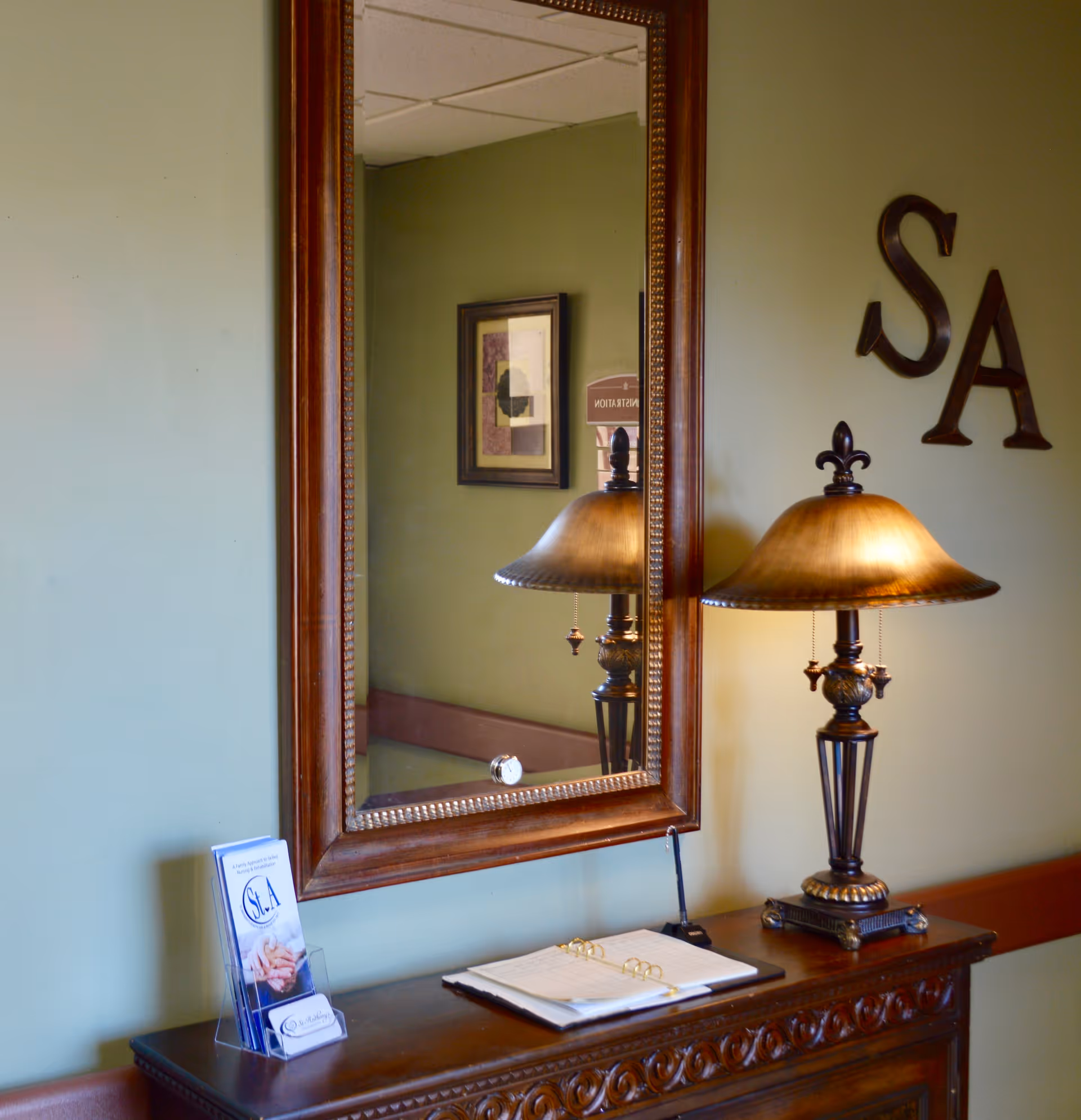 A wooden console table with intricate carvings against a green wall. On the table is a decorative lamp with a golden shade, an open binder, and a brochure holder containing brochures for St Anthony Community Care Center. Above the table is a large rectangular mirror with a wooden frame reflecting part of the room, including a framed picture and a sign. The letters 'S' and 'A' are mounted on the wall to the right of the mirror.