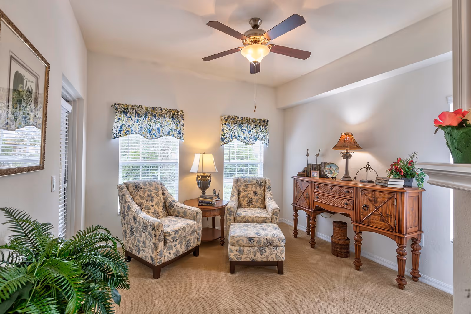 A cozy living room area with two floral-patterned armchairs and a matching ottoman arranged around a small round wooden side table with a lamp. Behind the chairs are two windows with floral valances. To the right is a wooden sideboard with decorative items and a table lamp. A ceiling fan with a light fixture is mounted on the ceiling, and a green potted plant is visible in the foreground.