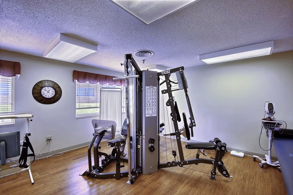 A fitness room with exercise equipment including a multi-station gym machine, stationary bike, and medical monitoring equipment. The room has wood flooring, white walls, two windows with red valances, a wall clock, and fluorescent ceiling lights.