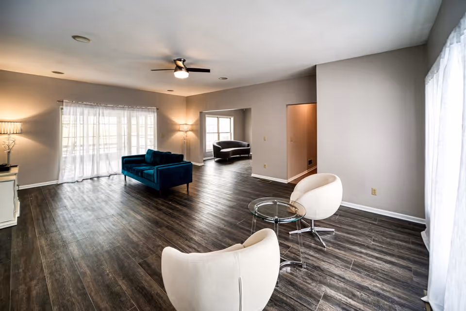 Spacious modern living room with dark wood-look floors, two white swivel chairs and a glass side table in the foreground and a teal sofa by large windows with sheer curtains.