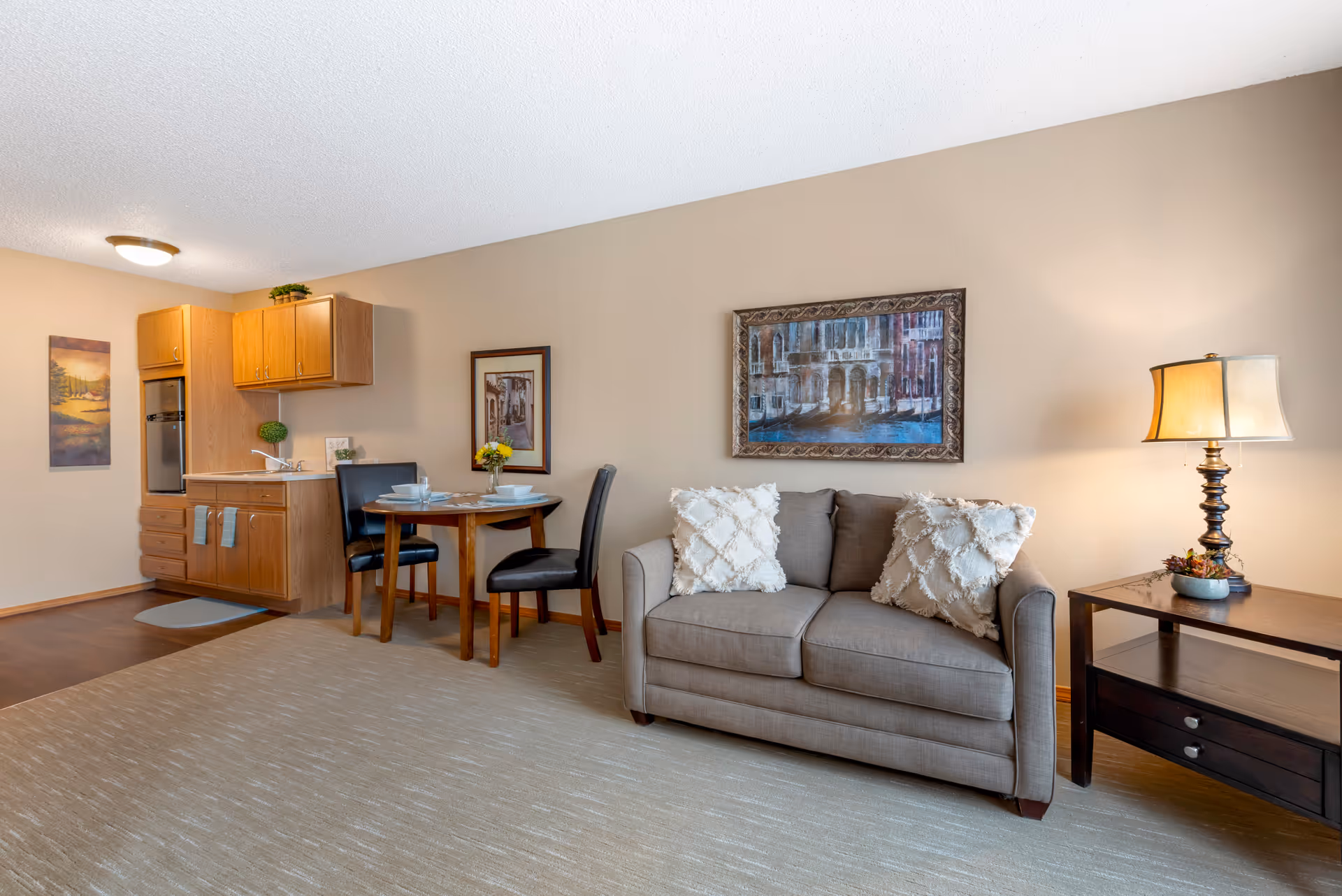 Living room and kitchenette area featuring a beige sofa with decorative pillows, a side table with lamp, a small dining table with chairs, and a compact kitchen.