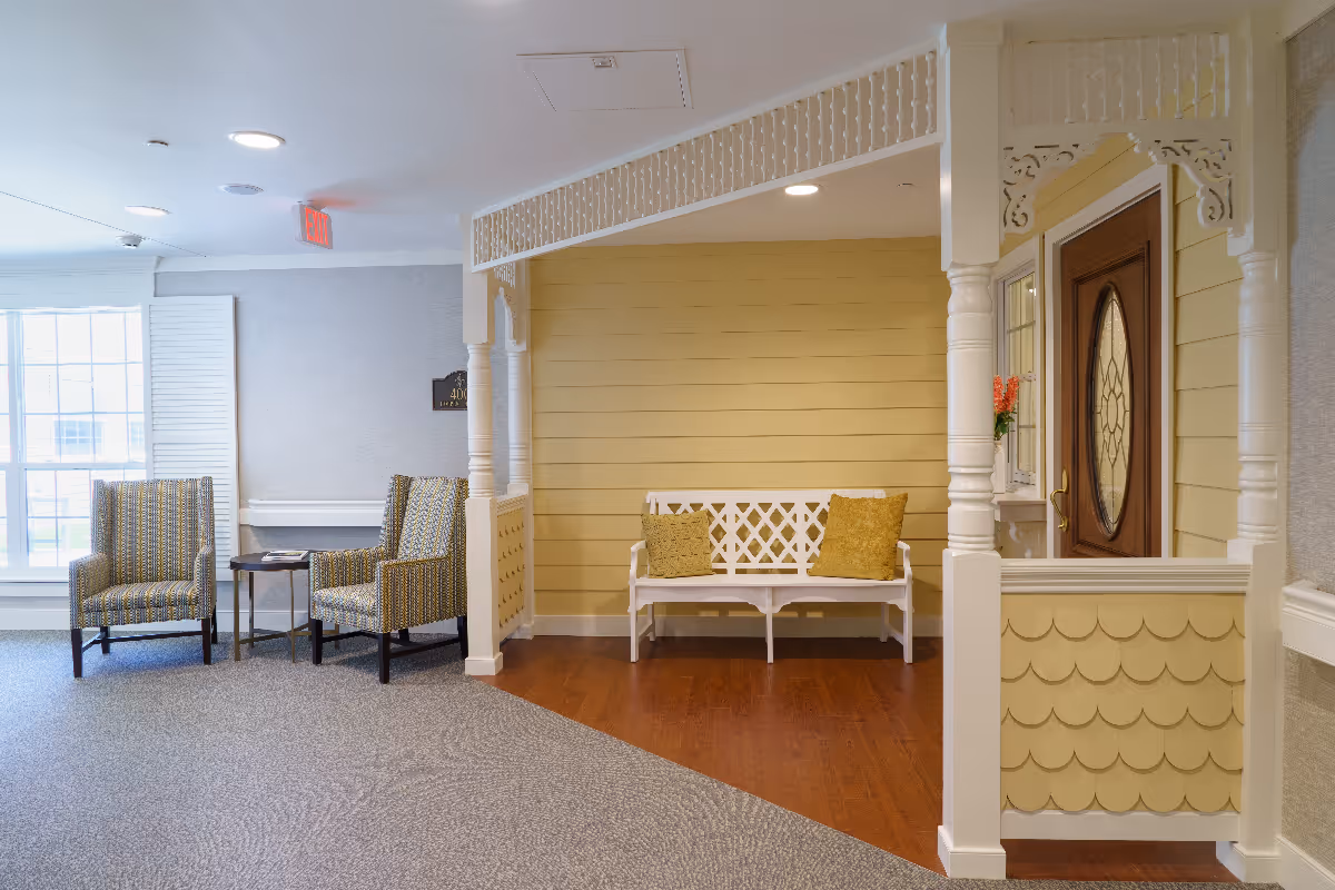 A cozy seating area in a senior living facility featuring two patterned armchairs with a small round table between them, a white wooden bench with two yellow cushions, decorative white columns and trim, a wooden door with an oval glass window, and a large window letting in natural light.