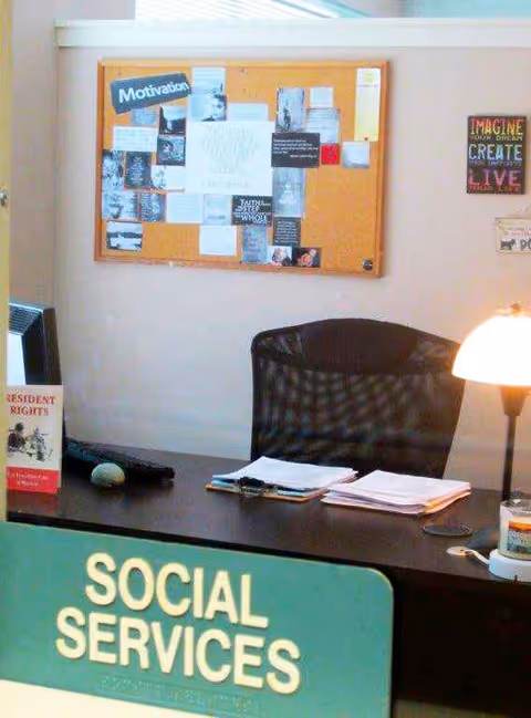 Office desk labeled Social Services with bulletin board, papers, lamp, and chair inside a facility.