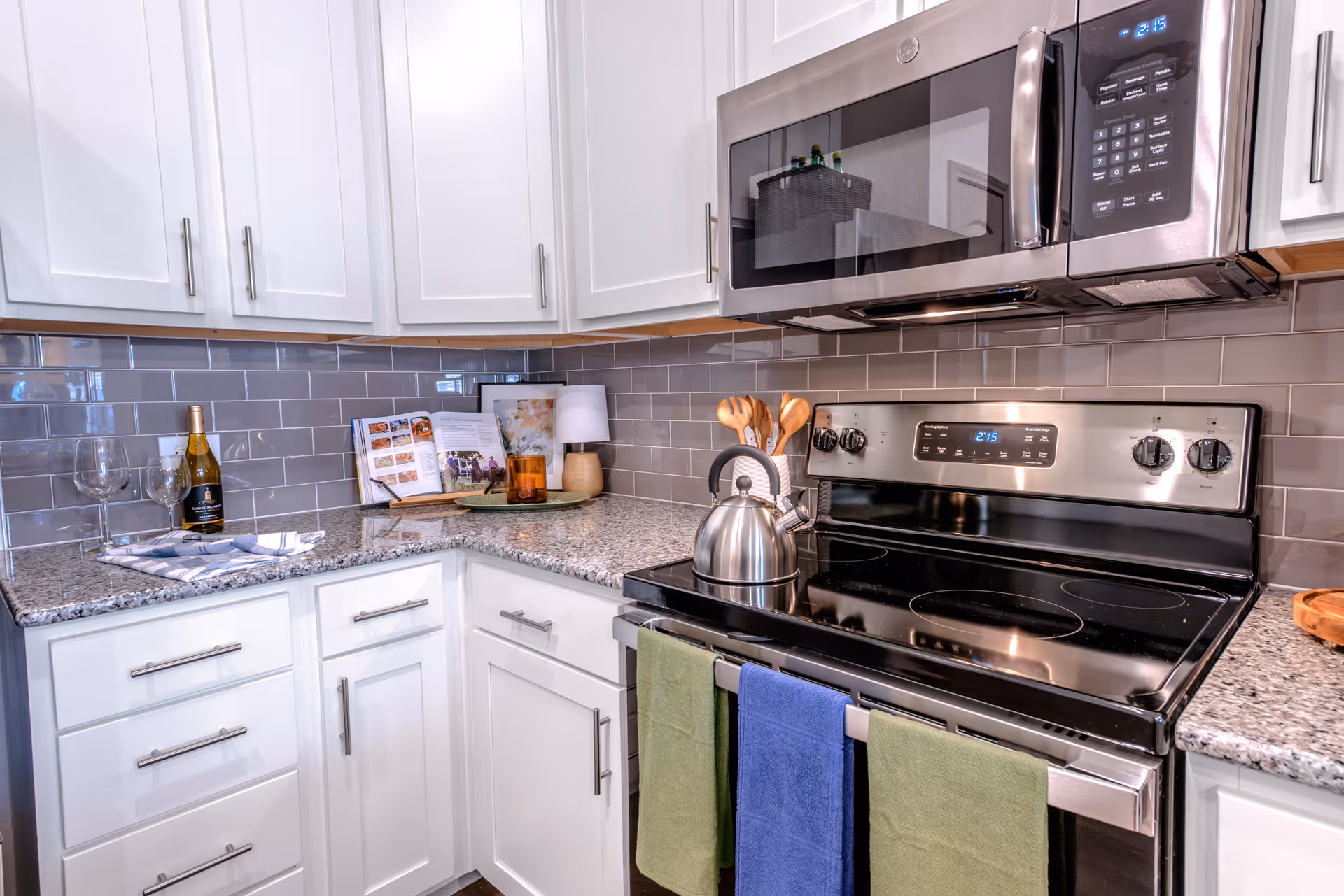 Modern kitchen corner with white cabinets, gray subway tile backsplash, granite countertops, stainless steel microwave and electric stove with a kettle on it. There are three towels hanging on the stove handle, a bottle of white wine with two wine glasses on the counter, a cookbook, a small lamp, and wooden cooking utensils in a white holder.