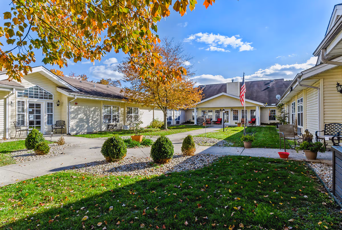 Outdoor courtyard area of Cedar Creek of Muncie facility with green grass, small bushes, a tree with autumn leaves, paved walkways, and beige buildings surrounding the courtyard under a blue sky with some clouds.