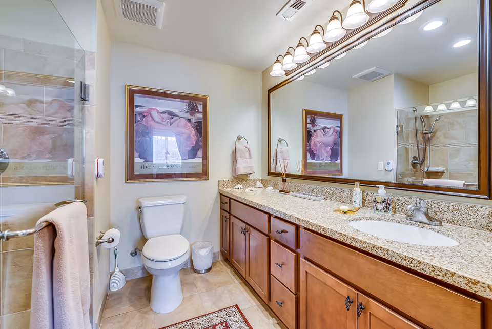 A clean and well-lit bathroom featuring a large mirror above a granite countertop with a sink, wooden cabinets underneath, a toilet, a glass-enclosed shower with beige tiles, a towel rack with a pink towel, and a framed artwork on the wall.