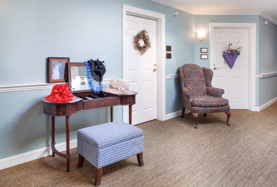 A hallway in a senior living facility with light blue walls and beige carpet. There is a wooden vanity table with a mirror and three decorative hats on it, along with a blue patterned cushioned stool. Next to the table is a patterned upholstered armchair. Two white doors are visible, each decorated with a wreath and a small sign beside them.