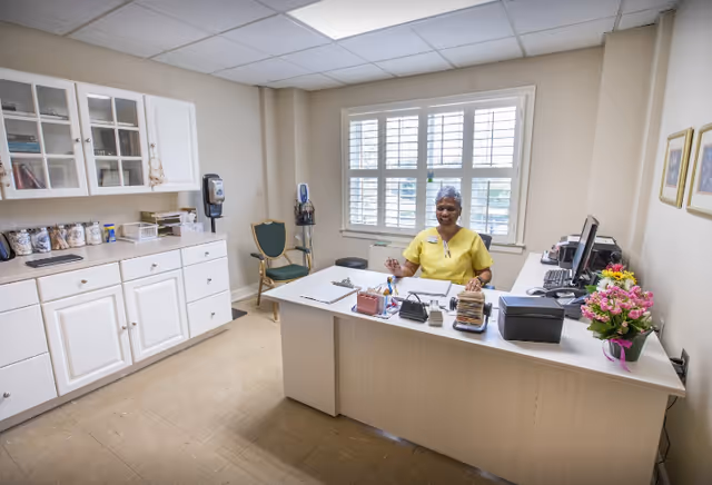 A smiling staff member in a yellow uniform sits behind a desk in a well-lit office room with a large window with white shutters. The desk is organized with office supplies, a computer, and a vase of pink flowers. White cabinets with glass doors and various items are visible on the left side of the room.
