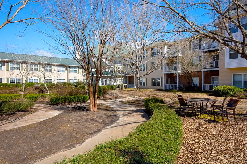 Sunny courtyard with paved walkways, leafless trees, a patio table and chairs, and surrounding multi-story assisted-living buildings.