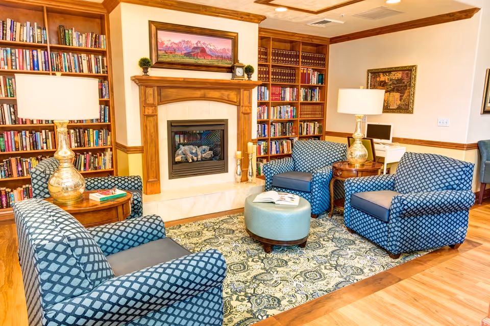 A cozy living room area with four blue patterned armchairs arranged around a small round ottoman with an open book on it. The room features a wooden fireplace with a painting above it, flanked by bookshelves filled with books. There are two side tables with decorative lamps, and a patterned area rug on the wooden floor. A small desk with a computer is visible in the background.