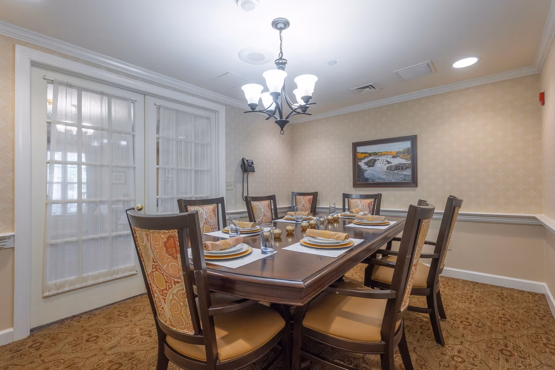 A dining room with a rectangular wooden table set for eight people. Each place setting includes a plate, napkin, and glass. The chairs have patterned upholstery. The room has beige patterned wallpaper, a carpeted floor, a chandelier overhead, and a framed painting of a river on the wall. French doors with sheer curtains are visible on one side.