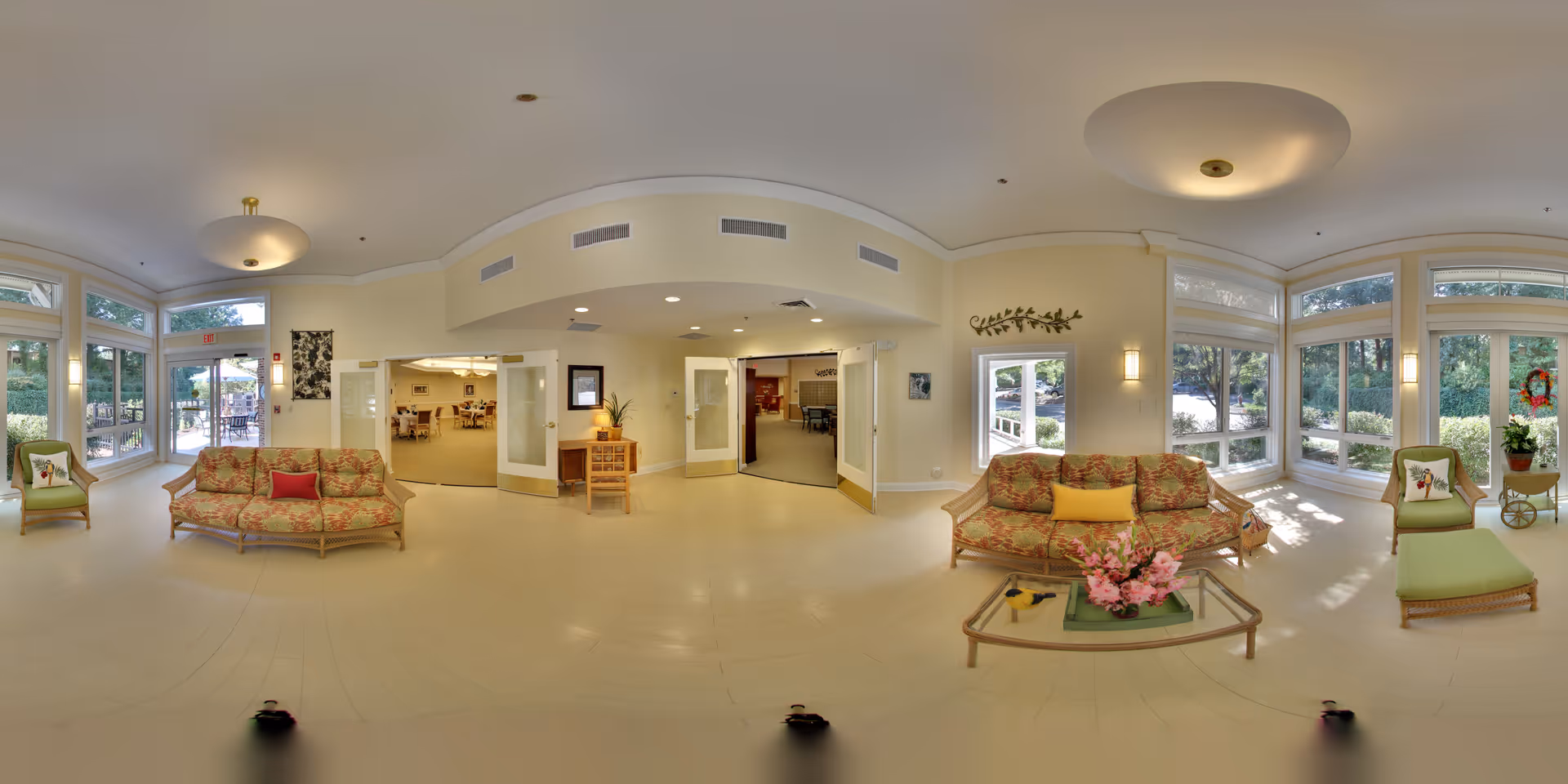 A spacious and bright common area in a senior living facility with large windows letting in natural light. The room features two floral-patterned sofas with colorful cushions, a glass coffee table with a pink flower arrangement, and several chairs with green cushions. There are double doors leading to other rooms, and the walls are painted a soft beige. Outside, greenery and trees are visible through the windows.