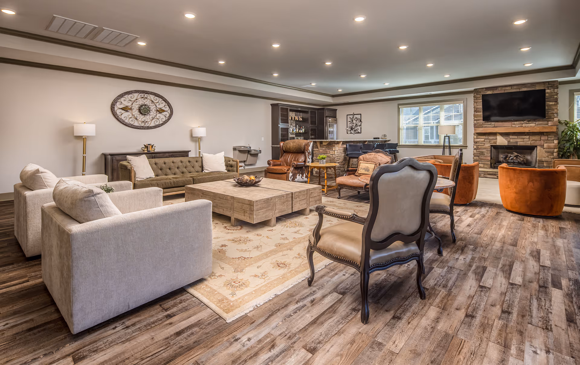 A spacious and well-lit living room with various seating options including beige sofas, a green tufted couch, brown leather and upholstered chairs, and orange armchairs. The room features a large wooden coffee table on a patterned rug, a stone fireplace with a mounted TV above it, a bar area with stools, and decorative wall art. The floor is wood with a natural finish, and there are multiple recessed ceiling lights.