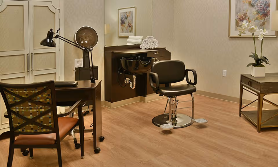 Interior view of a salon area in a senior living facility featuring a black salon chair in front of a black wash basin, a wooden desk with a black adjustable lamp, a chair with a patterned backrest, a side table with a white orchid plant, and framed floral artwork on the beige patterned walls.