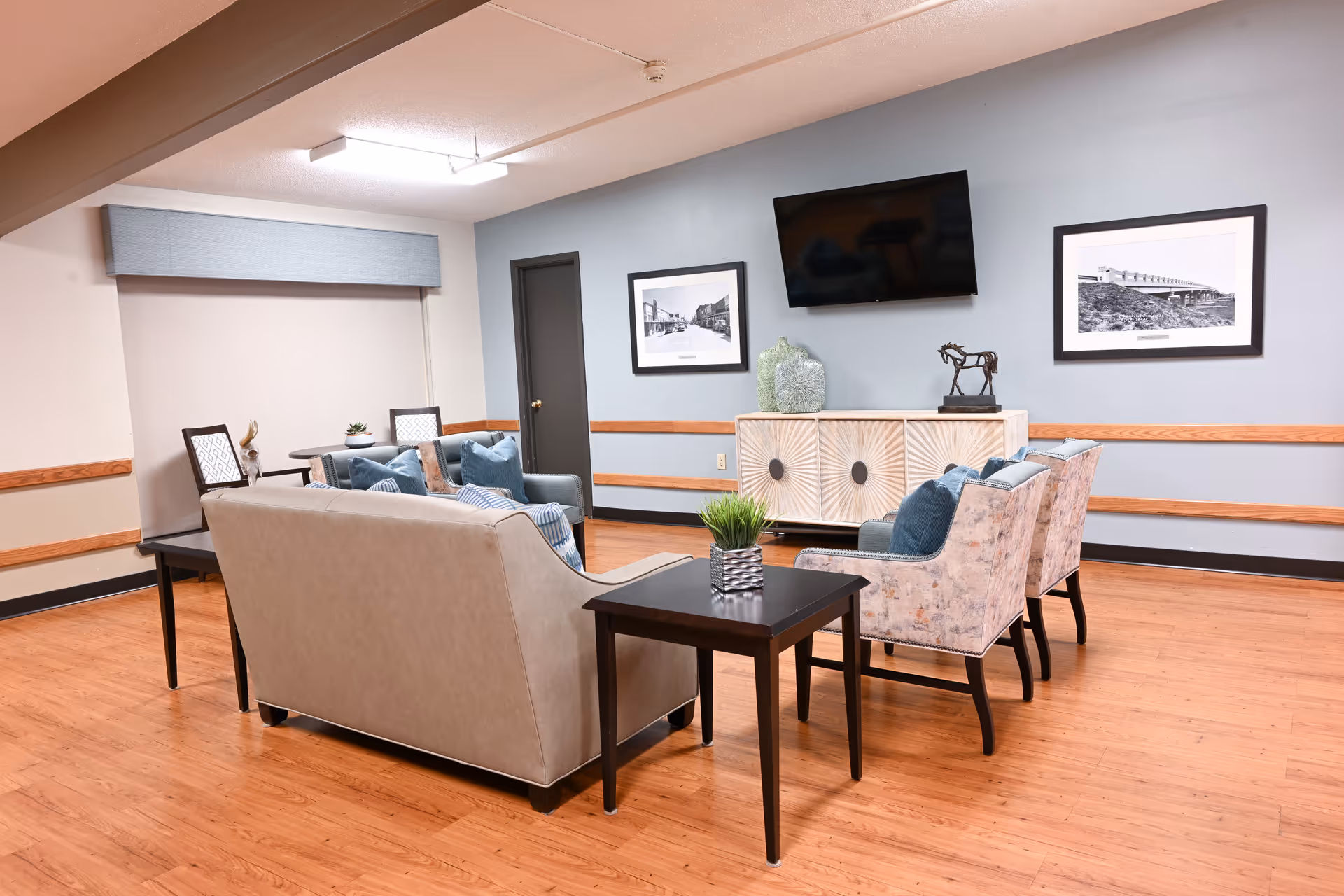 A cozy living room area in a nursing and rehabilitation center featuring a beige sofa, four patterned armchairs with blue cushions, two side tables with small plants, a white decorative cabinet, a wall-mounted flat-screen TV, and two black-and-white framed photographs on a light blue wall.