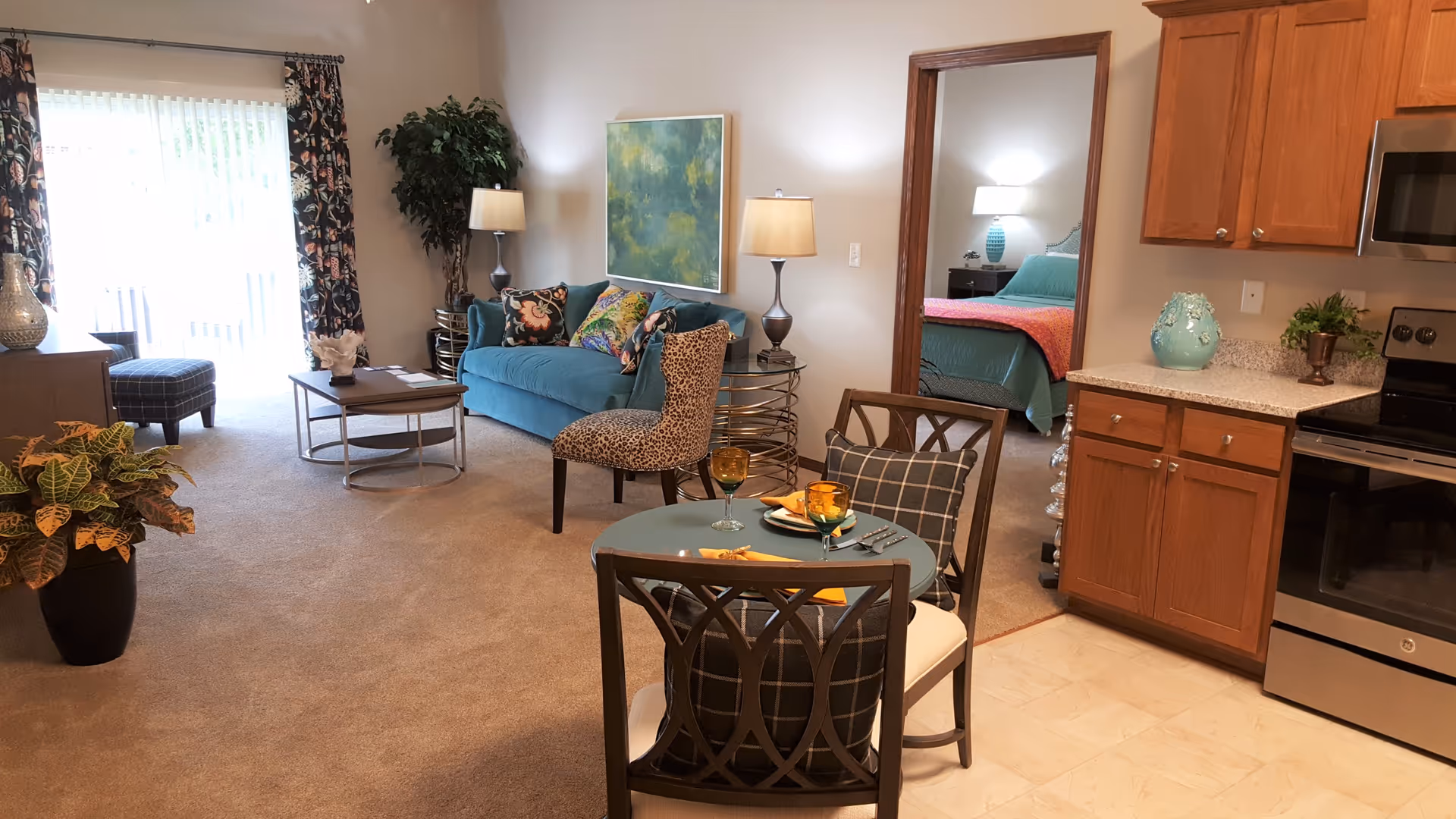 A cozy living space featuring a teal sofa with colorful pillows, a leopard print chair, two side tables with lamps, a small round dining table set for two with amber glasses and napkins, and a kitchen area with wooden cabinets and stainless steel appliances. A bedroom with a teal bedspread and a pink throw is visible through an open doorway. The room is carpeted with beige carpet and has a large window with floral curtains letting in natural light.