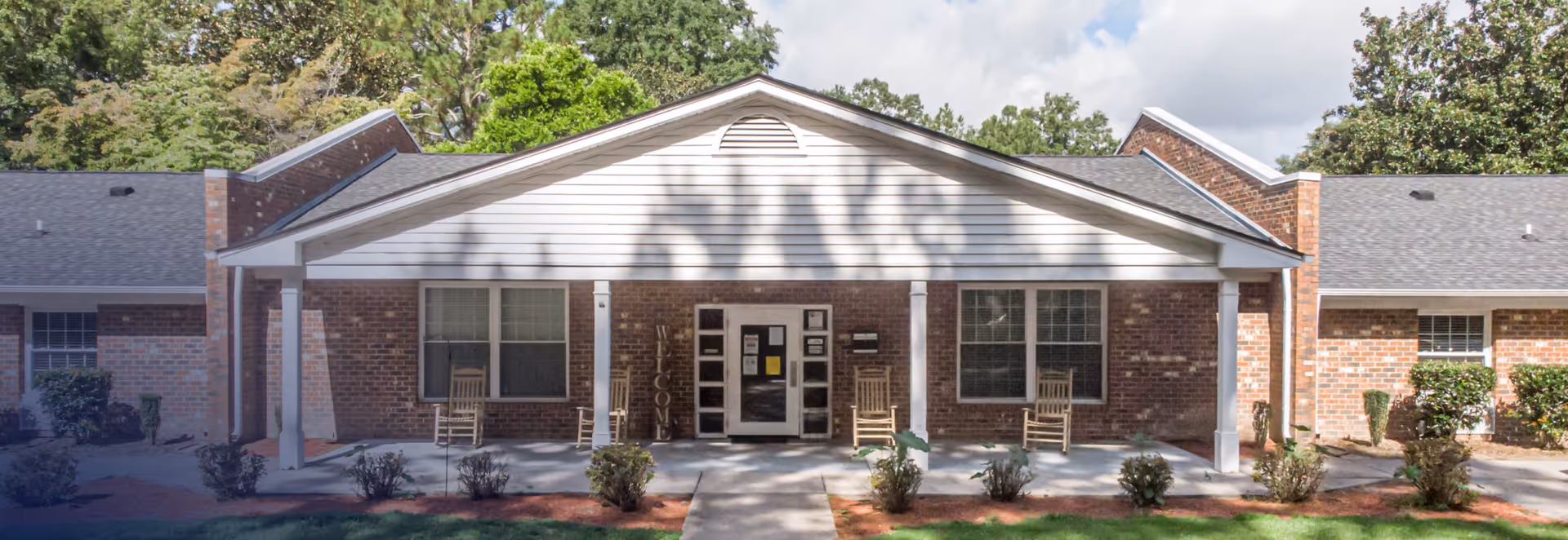 Front entrance of a single-story brick building with a covered porch, rocking chairs, and small landscaped shrubs.